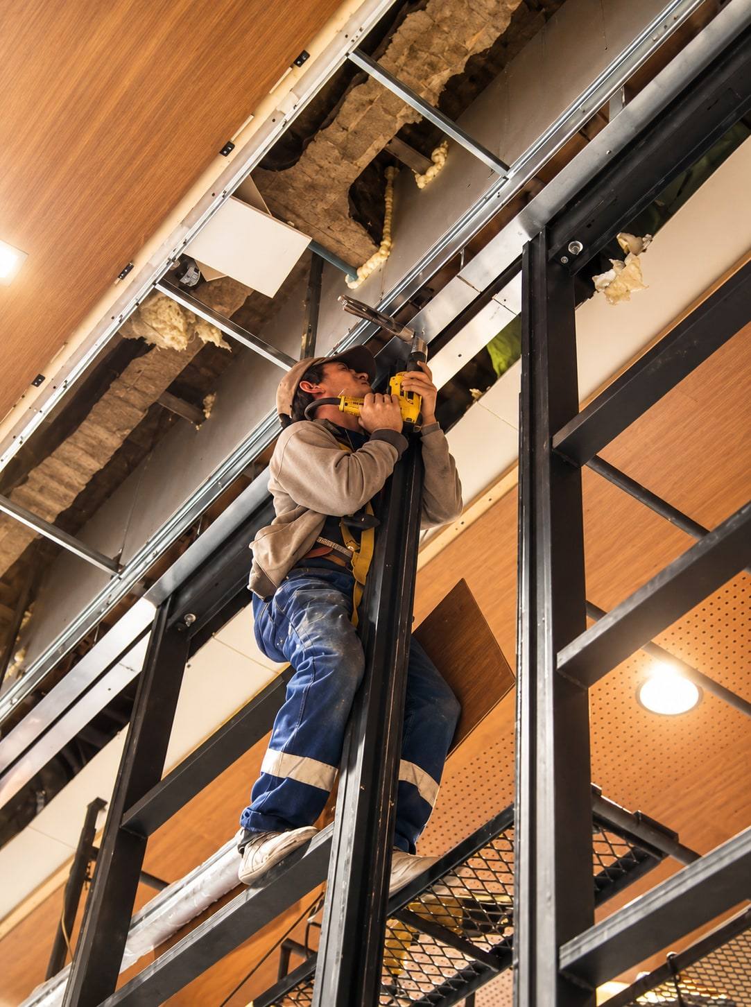 Instalador con taladro sobre una estructura metálica, bajo un techo con paneles de madera y aislante blanco.